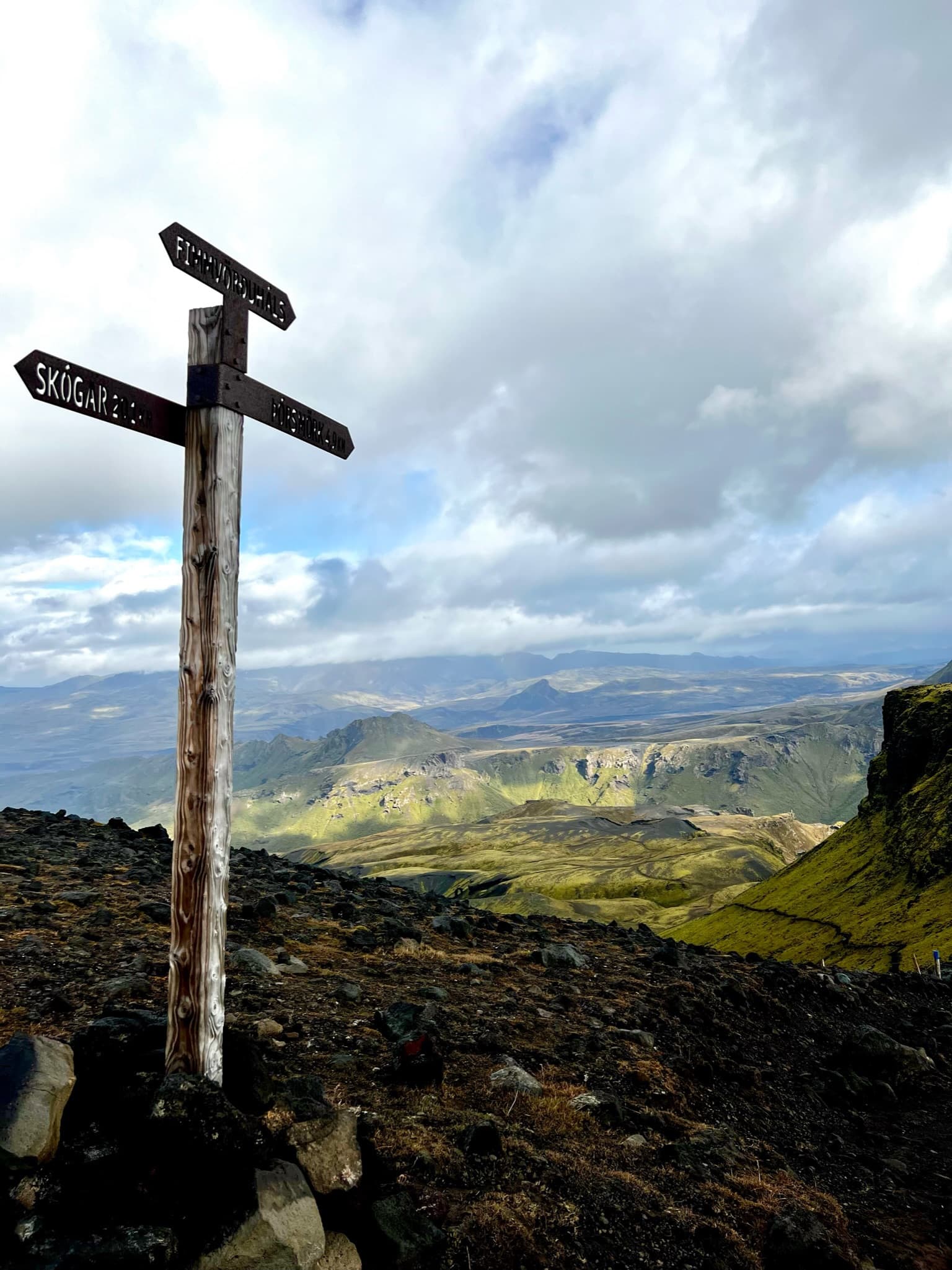 A weathered wooden trail signpost at the Fimmvörðuháls junction in Iceland, pointing toward Skógar and Þórsmörk above a green highland valley.